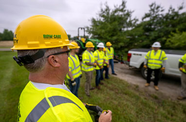A member of the Row-Care crew wears a branded yellow hard hat and stands among other team members in a field.
