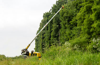 A yellow Row-Care trimmer with its white arm outstretched toward a line of trees in a field.