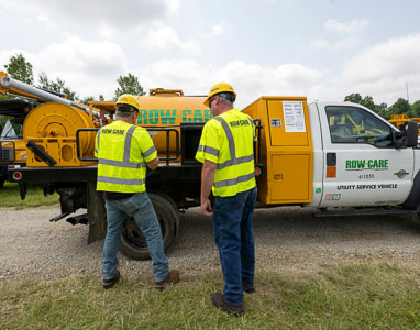 The backs of two Row-Care team members wearing neon yellow branded safety vests. They stand in front of a large yellow piece of Row-Care equipment.